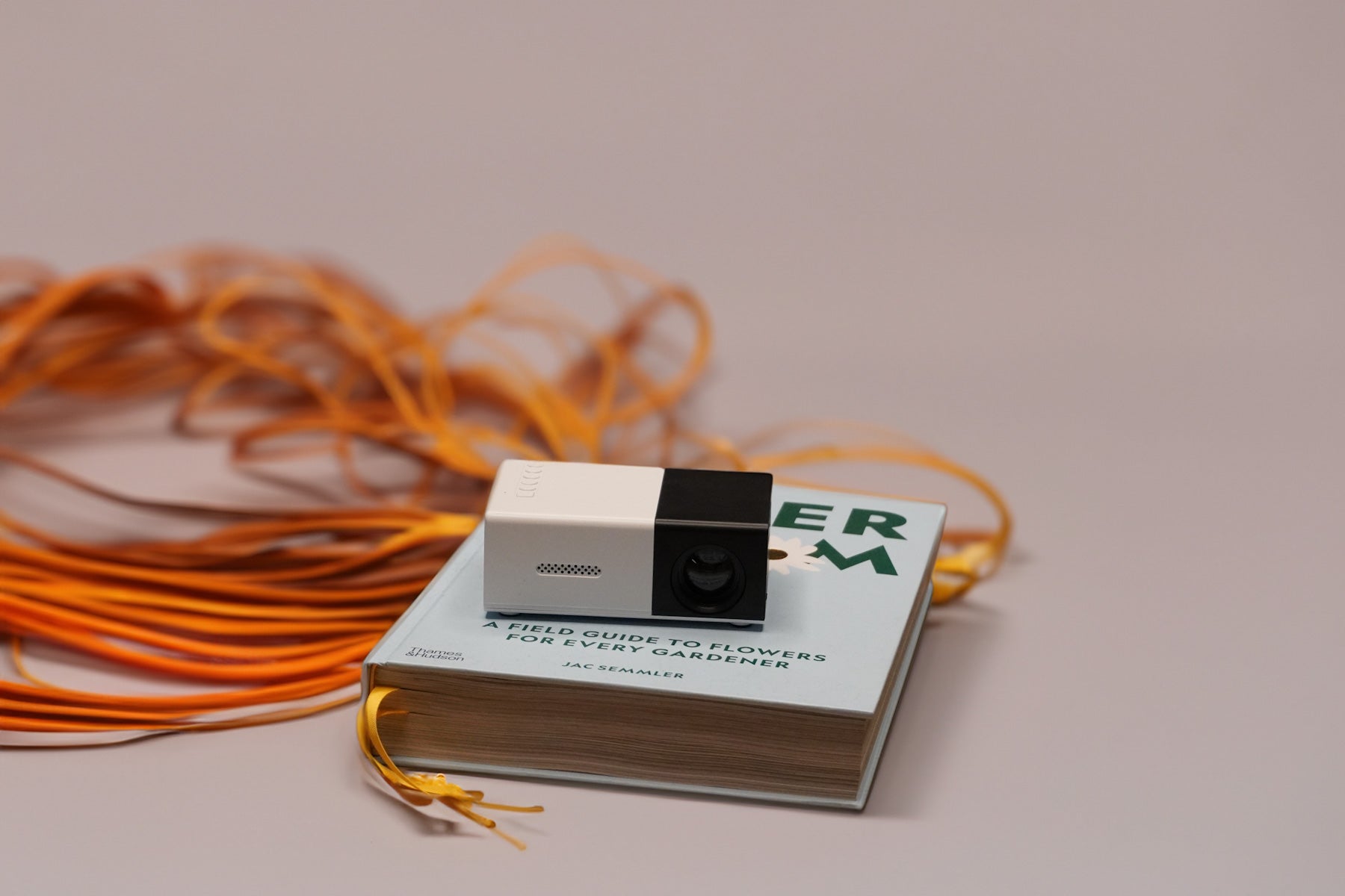 A small white and black projector sits on a closed book titled A Field Guide to Flowers for Every Gardener. Orange and yellow ribbons are spread out behind the book on a neutral background.