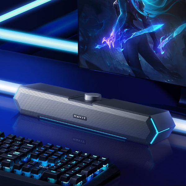 A stylish The Projection Room RGB Bluetooth Speakers with RGB lighting sits on a glossy desk in front of gaming speakers, keyboard, and monitor, featuring a handy 3.5MM audio jack for easy connectivity.