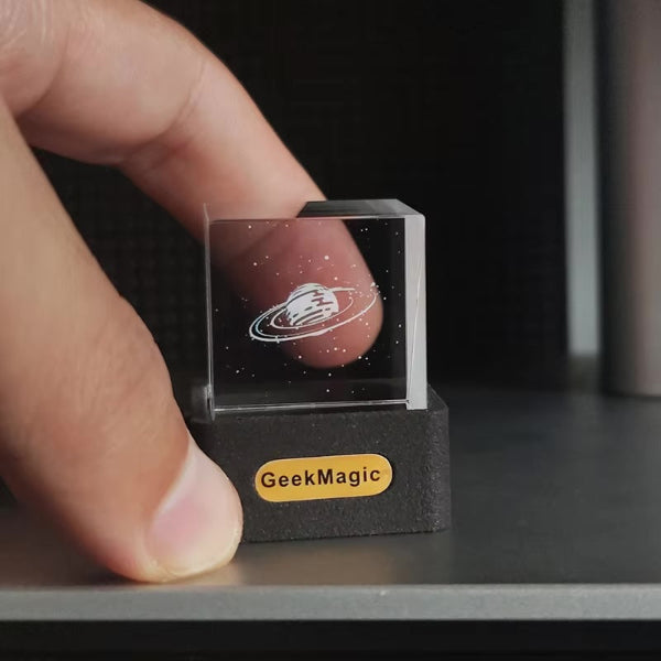 A hand holds a 3D Icon Desk Light Decor by The Projection Room—a small glass cube engraved with Saturn and stars, resting on a black GeekMagic base. Connect it to your mobile app for customizable control, adding magic to your space.