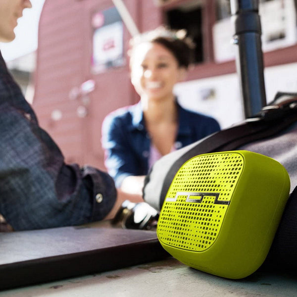 A SOL Republic Punk Wireless Pocket Speaker by The Projection Room, small and square in lime-green, sits on a table beside bags. In the blurred background, a dark-haired person in a denim shirt smiles, perhaps chatting with another person partly visible.
