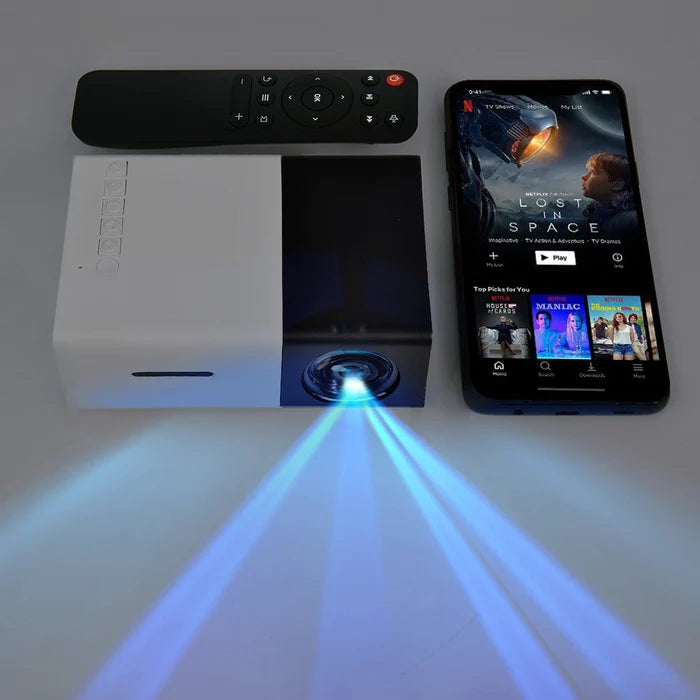 A compact projector beams blue light next to a smartphone displaying a streaming app and a black remote control, all placed on a gray surface.