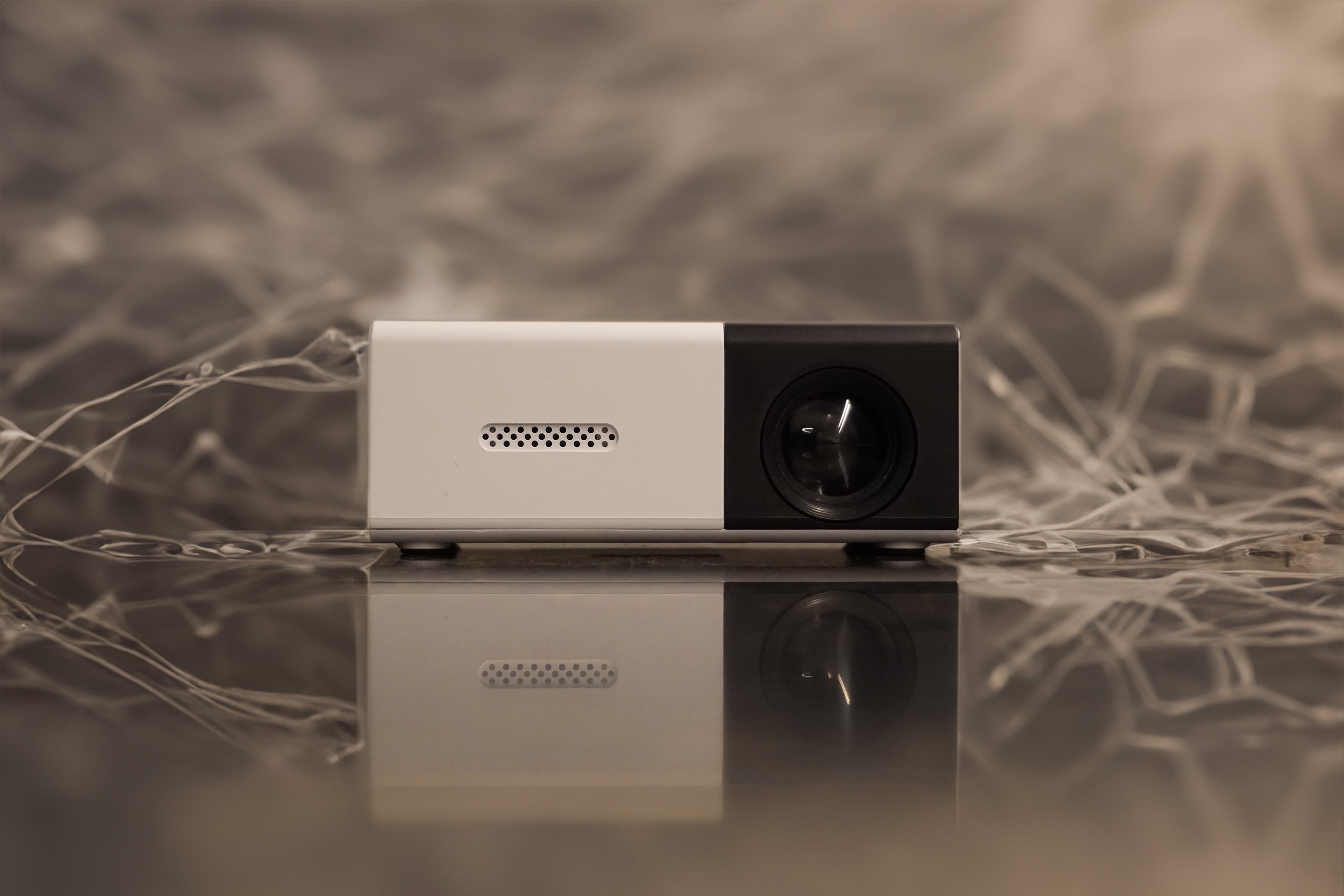A small, modern projector is placed on a reflective surface with an abstract, web-like background pattern, creating a symmetrical visual effect.