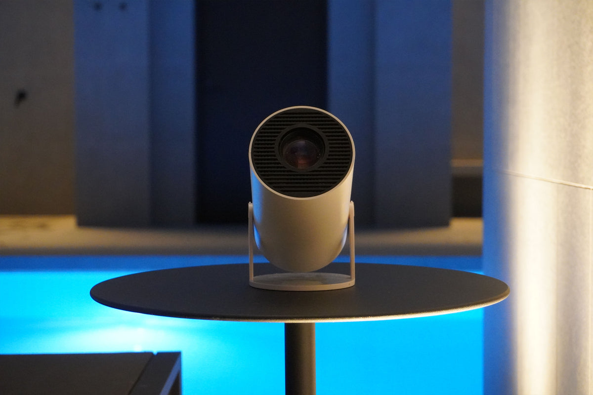 A cylindrical projector sits on a round black table, facing forward. The background is softly lit with shades of blue and gray, creating a modern, ambient setting.