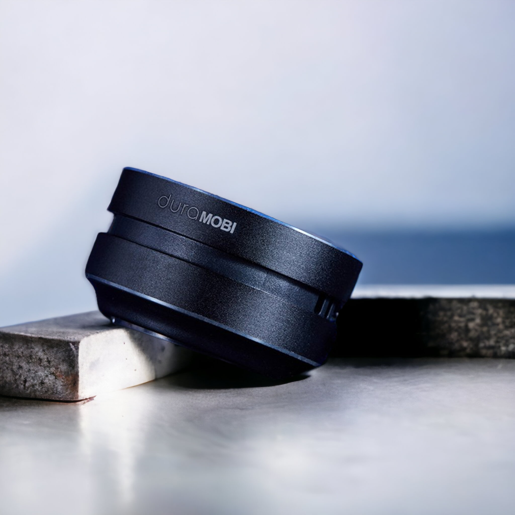 A black duroMOBI camera lens attachment rests at an angle on two marble slabs against a blurred, neutral background.