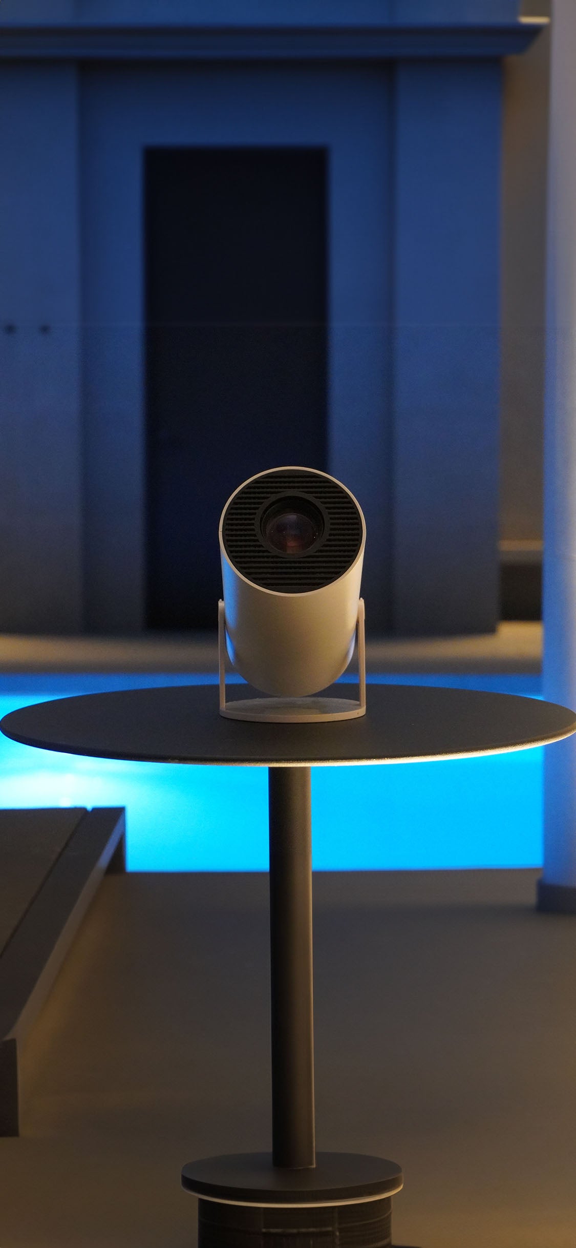 A modern cylindrical projector sits on a small round table in a dimly lit room with blue lighting and a dark doorway in the background.