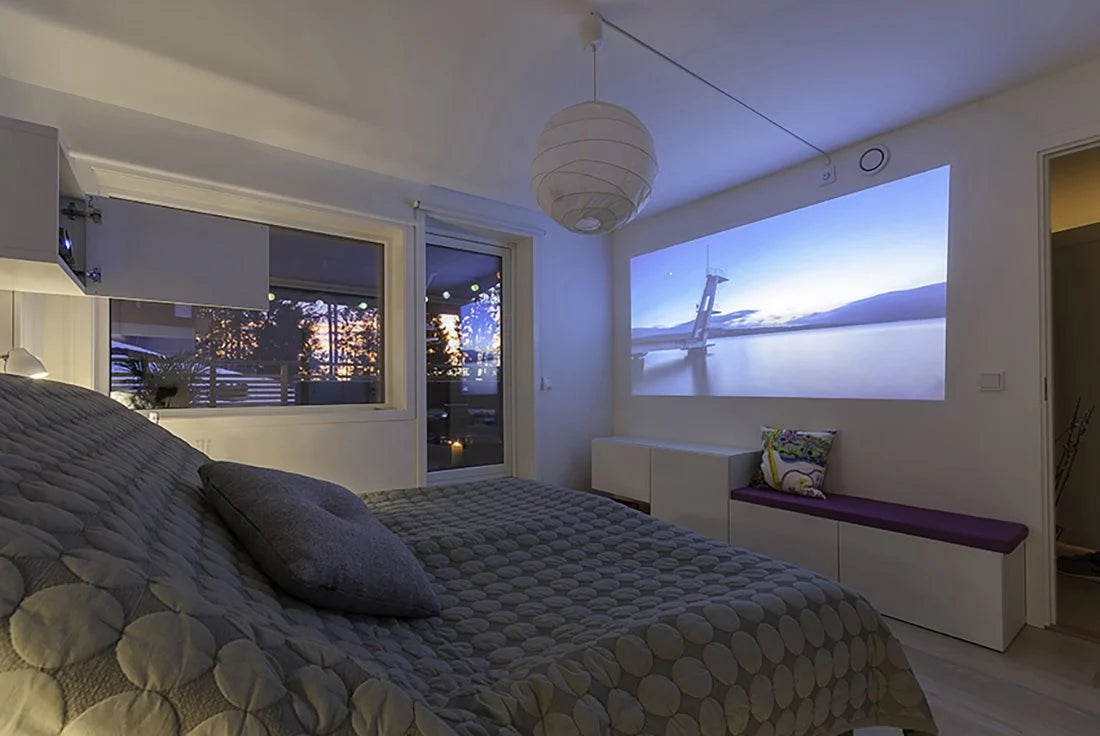A modern bedroom with a patterned bedspread, large windows, and a projector displaying a scenic landscape on the wall. There is a white pendant lamp above and a bench with a purple cushion by the window.